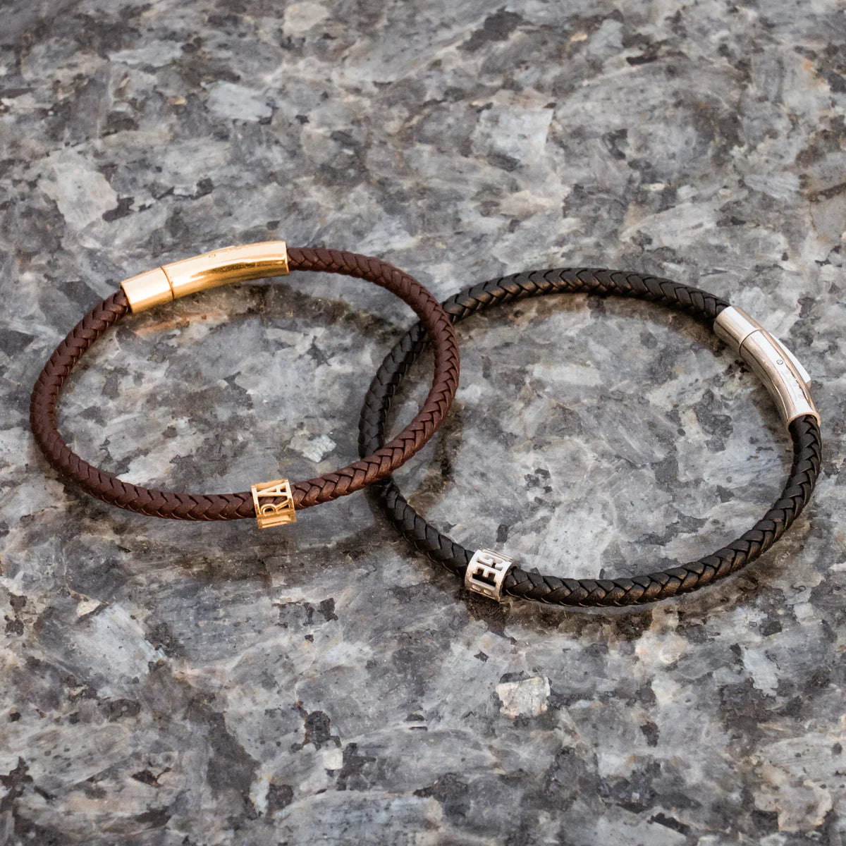 Two braided leather bracelets with gold and silver metal ends on a gray stone surface