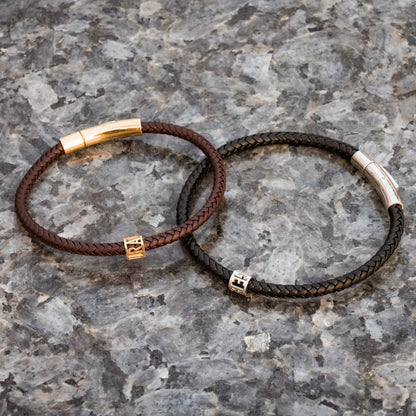 Two braided leather bracelets with gold and silver metal ends on a gray stone surface