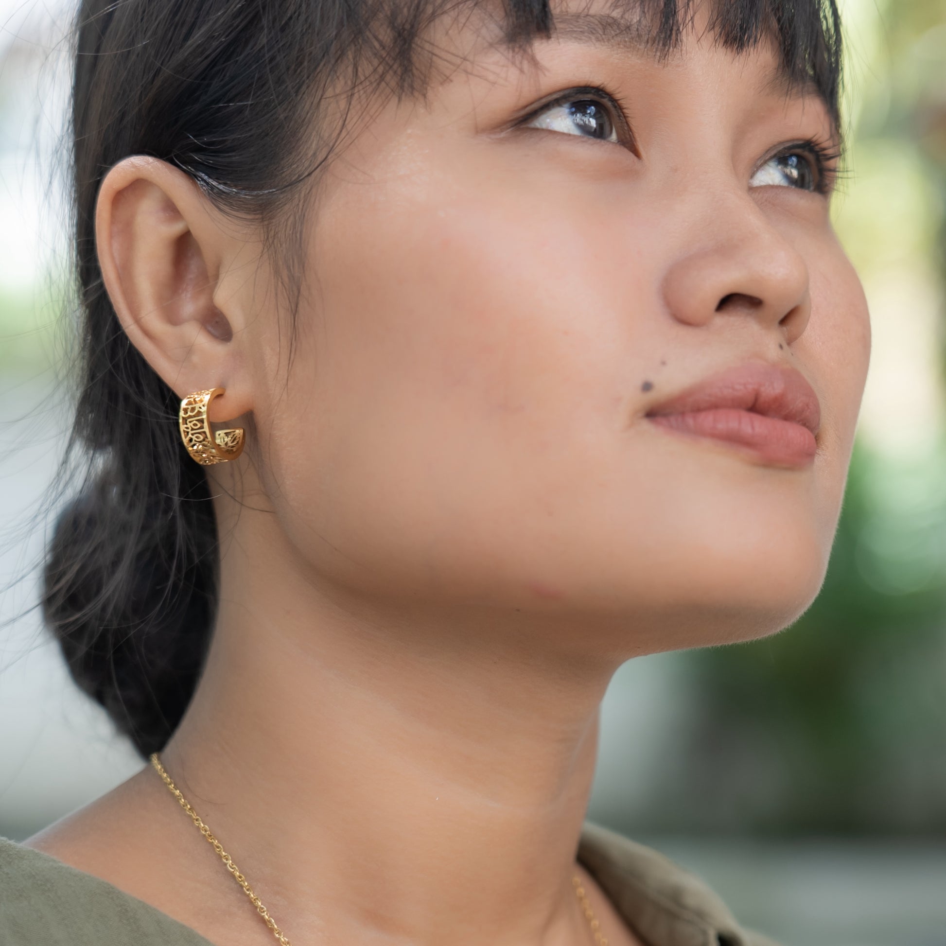 Close-up of a woman wearing gold earrings and a necklace with a blurred natural background