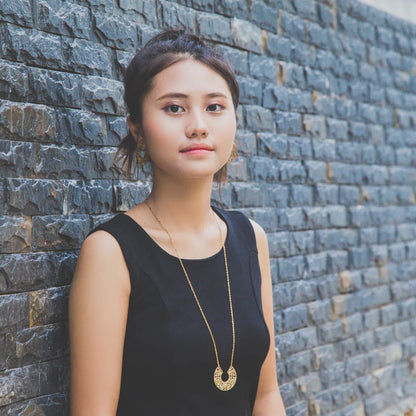 Woman wearing a black sleeveless top and gold necklace against a stone wall.