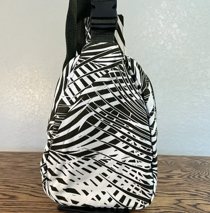 A black and white abstract patterned sling bag with a black shoulder strap, standing on a wooden surface against a light gray wall.