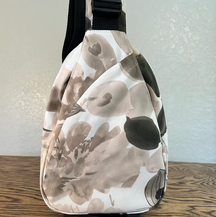 A slouchy shoulder bag with a watercolor floral pattern in beige, brown, and black on a white background, resting on a wooden surface against a textured wall.