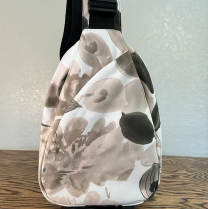 A slouchy shoulder bag with a watercolor floral pattern in beige, brown, and black on a white background, resting on a wooden surface against a textured wall.