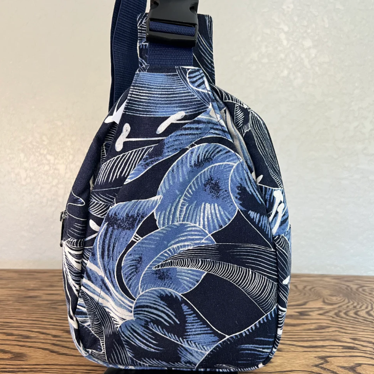 A navy blue fabric bag with a white tropical leaf pattern, featuring a black plastic buckle on the strap, standing on a wooden surface against a textured off-white wall.