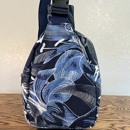 A navy blue fabric bag with a white tropical leaf pattern, featuring a black plastic buckle on the strap, standing on a wooden surface against a textured off-white wall.