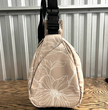 A beige sling bag with a white floral line pattern and a black adjustable strap, standing on a wooden surface against a corrugated metal wall.