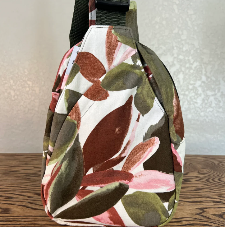 A fabric shoulder bag with a botanical leaf pattern in olive green, terracotta brown, and dusty pink on a white background, resting on a wooden surface against a textured light gray wall.