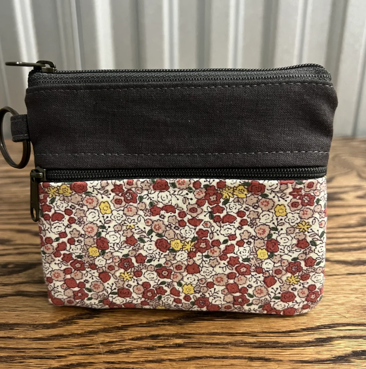 A small zip-around coin pouch with a solid black top section and a floral patterned bottom section, shown on a wooden tabletop with a white paneled background.