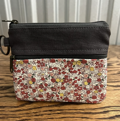 A small zip-around coin pouch with a solid black top section and a floral patterned bottom section, shown on a wooden tabletop with a white paneled background.