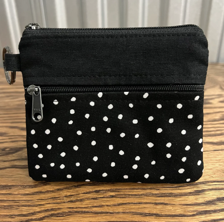 A small black fabric zip pouch with white polka dots on the lower half, shown on a wooden tabletop with a neutral indoor background.