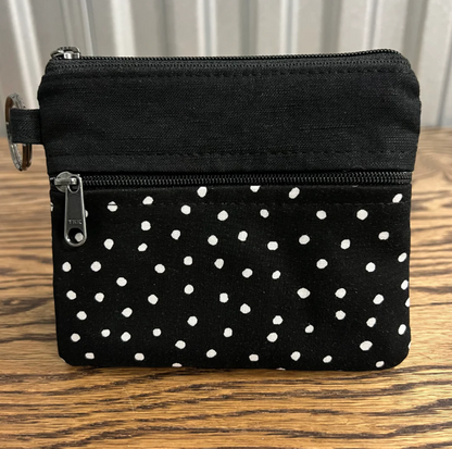 A small black fabric zip pouch with white polka dots on the lower half, shown on a wooden tabletop with a neutral indoor background.