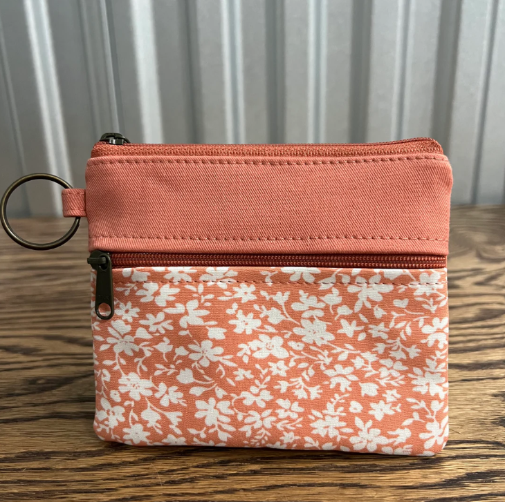A small zippered pouch with a coral pink top panel and a white floral print lower section, resting on a wooden surface in front of a gray curtain background.
