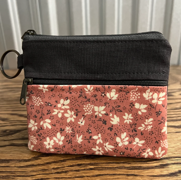 A small zippered pouch with a two-tone design: a solid black top section and a dusty rose pink bottom section with a white floral print. It has a black metal keyring attached to the left zipper pull and is photographed on a wooden tabletop with a neutral background.