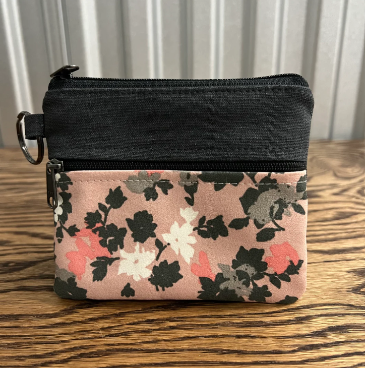 A small zippered pouch with a black top section and a pink floral patterned bottom section, placed on a wooden surface in front of a white paneled background.