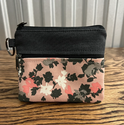 A small zippered pouch with a black top section and a pink floral patterned bottom section, placed on a wooden surface in front of a white paneled background.