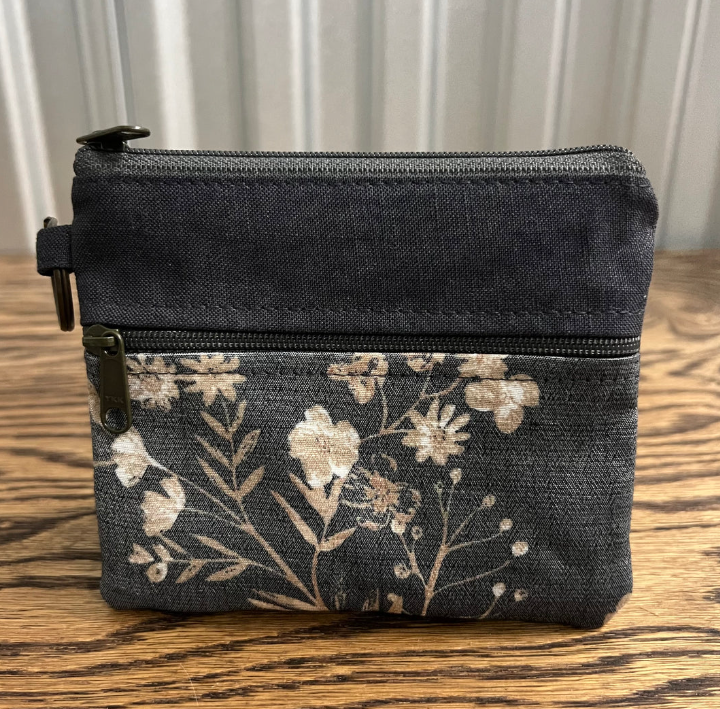 A small zippered pouch or coin purse made of dark charcoal gray fabric, featuring a floral print in beige and cream on the lower half. The pouch is shown resting on a wooden surface with a light-colored paneled wall in the background.