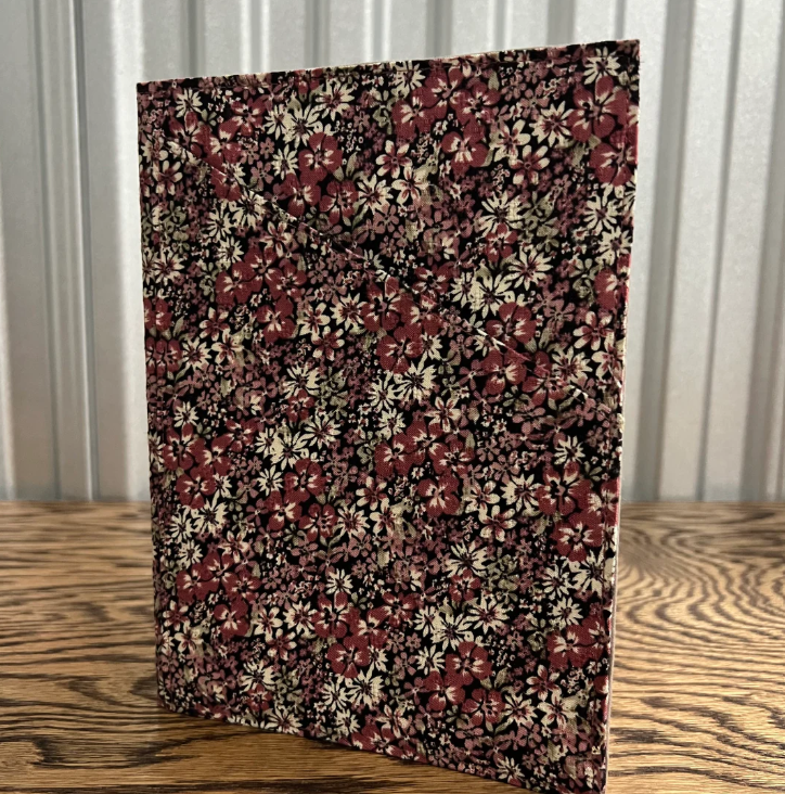 A rectangular fabric-covered notebook or folder with a dense floral print, standing upright on a wooden tabletop in front of a corrugated metal wall.