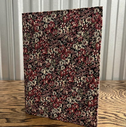 A rectangular fabric-covered notebook or folder with a dense floral print, standing upright on a wooden tabletop in front of a corrugated metal wall.