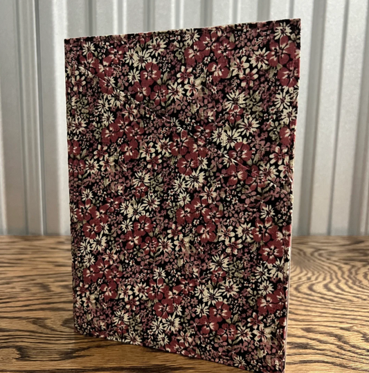 A rectangular fabric-covered notebook or folder with a dense floral print, standing upright on a wooden tabletop in front of a corrugated metal wall.