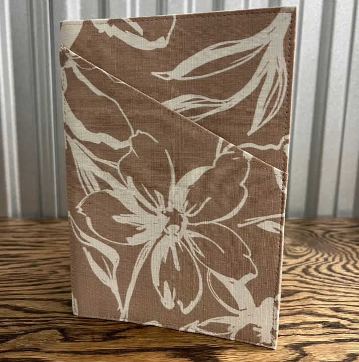 A fabric-covered notebook or journal with a brown and cream floral pattern, standing on a wooden surface against a corrugated metal background.