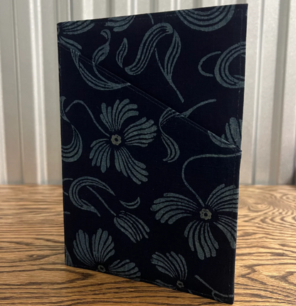 A black fabric sleeve or pouch with a gray floral pattern, standing on a wooden table in front of a white paneled background.