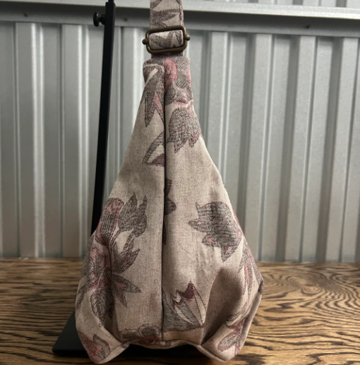 A slouchy shoulder bag made of beige linen fabric with a muted brown and dusty rose botanical pattern, displayed on a black stand against a corrugated metal wall and wooden floor.