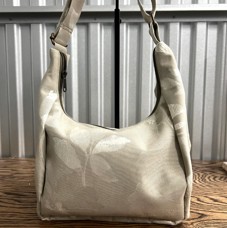 A beige shoulder bag with a subtle tonal leaf pattern, resting on a wooden surface in front of a corrugated metal wall.