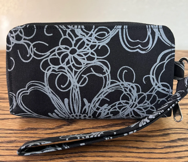 A black zip-around wallet with a white floral and scroll pattern, featuring a matching wristlet strap, resting on a wooden surface.