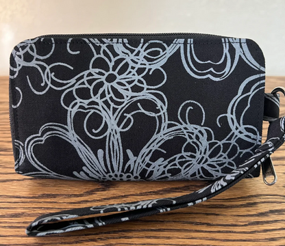 A black zip-around wallet with a white floral and scroll pattern, featuring a matching wristlet strap, resting on a wooden surface.