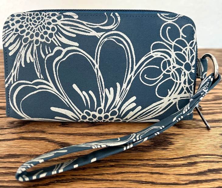 A dark blue fabric clutch wallet with a white floral and scroll pattern, featuring a matching wrist strap and metal ring, displayed on a wooden surface.