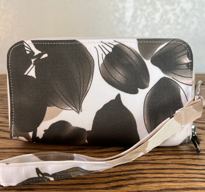 A zip-around wristlet wallet with a white background and a black floral tulip pattern, featuring a matching fabric wrist strap and a silver zipper pull, resting on a wooden surface.