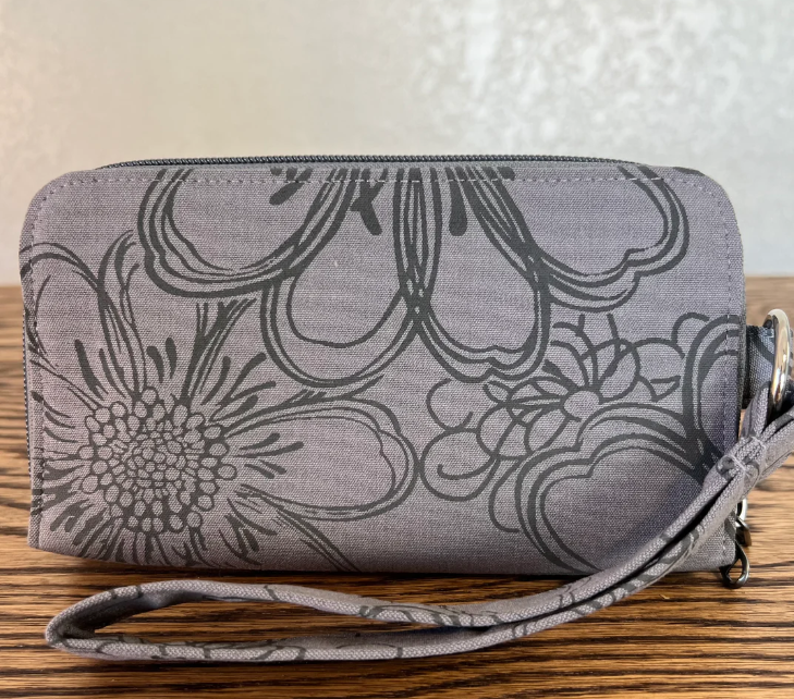A gray fabric zippered pouch with a black floral line-art pattern and a matching wrist strap, resting on a wooden surface against a light textured wall.