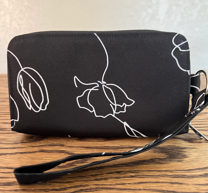 A black zip-around wristlet pouch with a white minimalist line-art floral pattern, resting on a wooden tabletop.