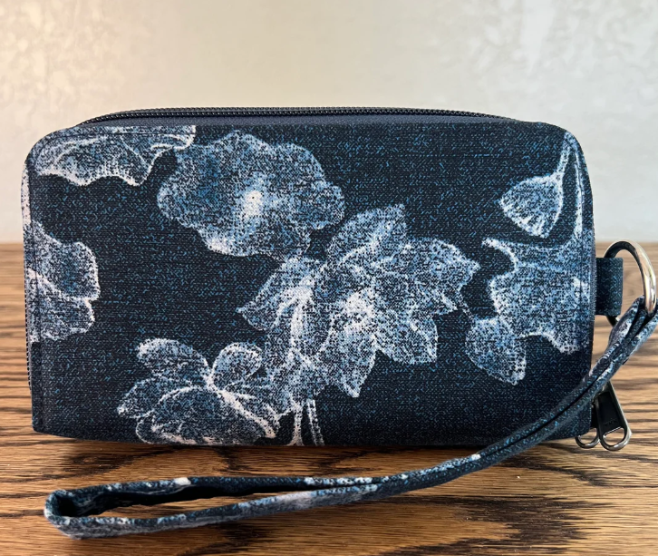 A rectangular zippered pouch or clutch in a dark denim-like fabric with a white floral print, shown on a wooden surface against a light beige wall. A matching wrist strap is attached on the right side.