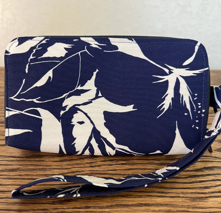 A zippered wristlet clutch with a navy blue and off-white tropical floral print, featuring a matching wrist strap, resting on a wooden surface.