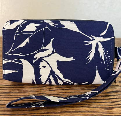 A zippered wristlet clutch with a navy blue and off-white tropical floral print, featuring a matching wrist strap, resting on a wooden surface.