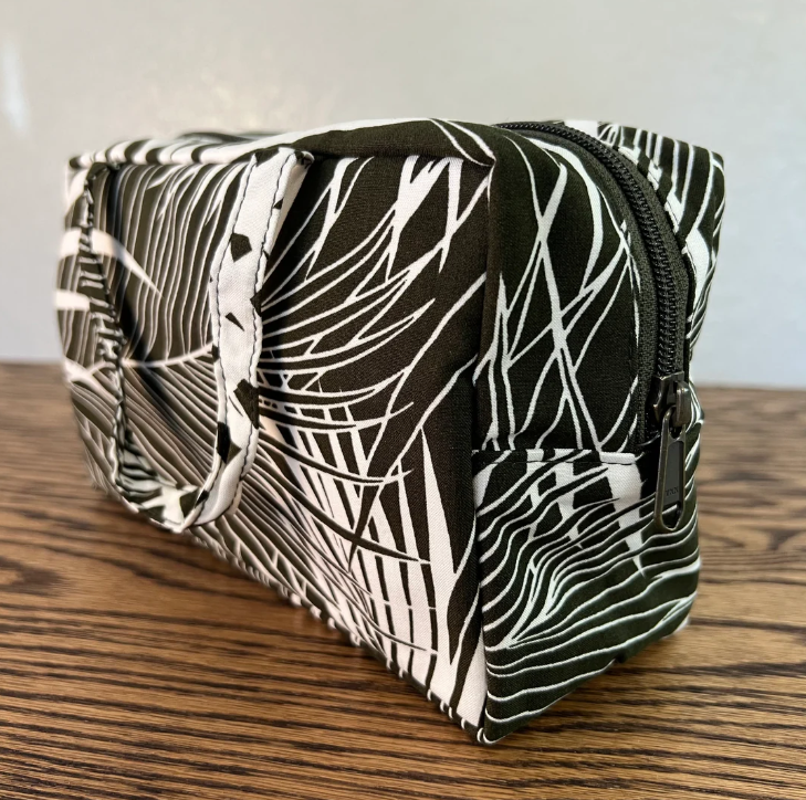 A fabric zippered pouch with a black and white abstract palm leaf pattern, resting on a wooden tabletop against a light gray wall.