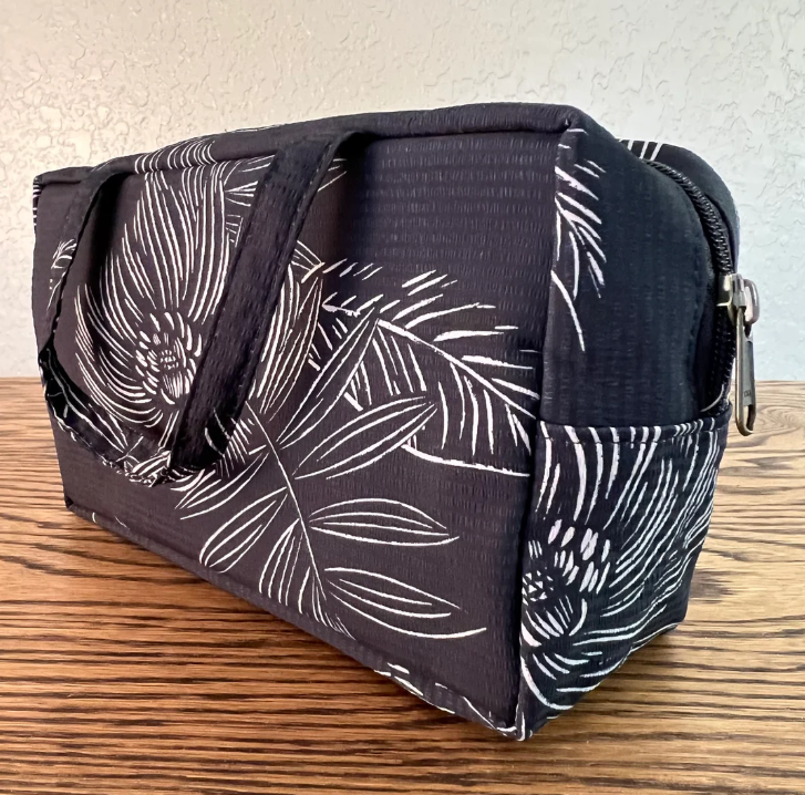 A fabric zippered pouch or small bag with a dark charcoal background and white tropical leaf and floral print, shown on a wooden surface against a light textured wall.