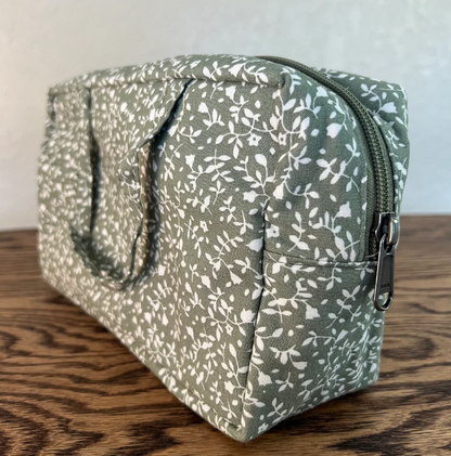 A fabric zippered toiletry bag with a white floral pattern on a sage green background, resting on a wooden surface against a light wall.