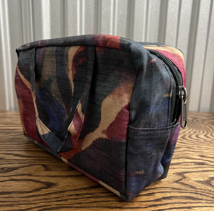 A rectangular zippered pouch with a dark abstract watercolor pattern in navy, burgundy, and beige, resting on a wooden tabletop against a corrugated metal background.