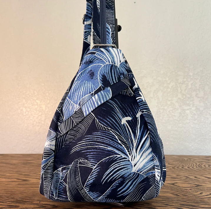 A slouchy shoulder bag with a tropical leaf print in navy blue, light blue, and white, resting on a wooden surface against a plain beige wall.