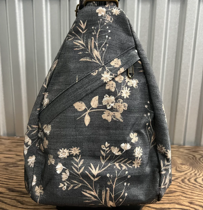 A dark gray fabric bag with a beige floral and botanical pattern, resting on a wooden surface against a corrugated metal background.