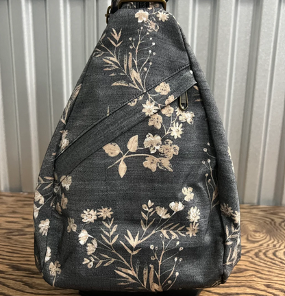 A dark gray fabric bag with a beige floral and botanical pattern, resting on a wooden surface against a corrugated metal background.