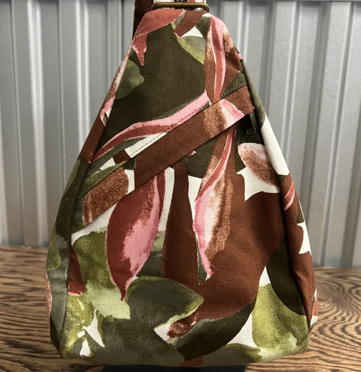 A slouchy shoulder bag with a bold abstract floral pattern in brown, pink, olive green, and white, resting on a wooden surface against a corrugated metal background.
