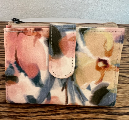 A small zip-around wallet with a floral watercolor print in pink, beige, blue, and brown tones, featuring a snap tab closure on the front, resting on a wooden surface.
