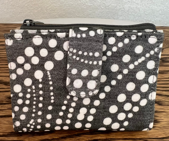 A fabric zippered pouch with an allover white polka dot pattern on a dark charcoal gray background, resting on a wooden surface against a light textured wall.
