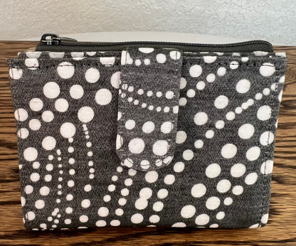 A fabric zippered pouch with an allover white polka dot pattern on a dark charcoal gray background, resting on a wooden surface against a light textured wall.