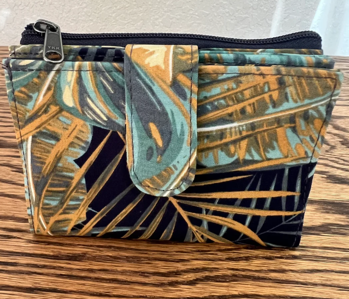 A small zippered pouch with a tropical palm leaf print in teal, gold, and black, resting on a wooden surface.