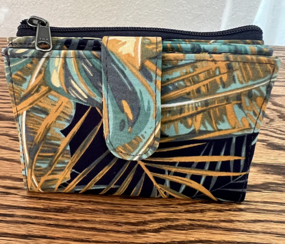A small zippered pouch with a tropical palm leaf print in teal, gold, and black, resting on a wooden surface.