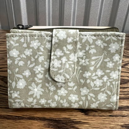 A small zippered fabric pouch or wallet with a white floral print on a light sage green background, featuring a stitched tab closure on the front, resting on a wooden surface.
